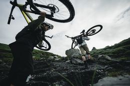 zwei Mountainbiker tragen ihre Räder über felsiges Gelände unter bewölktem Himmel
